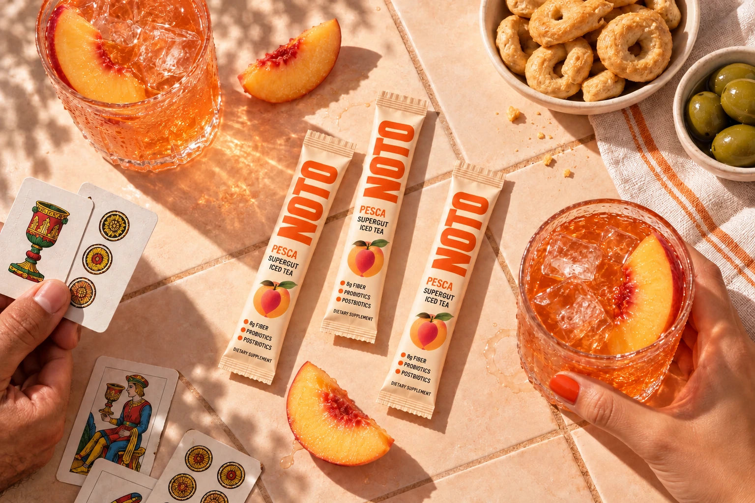 NOTO peach iced tea and sachets on a sunlit table with cards and aperitif snacks